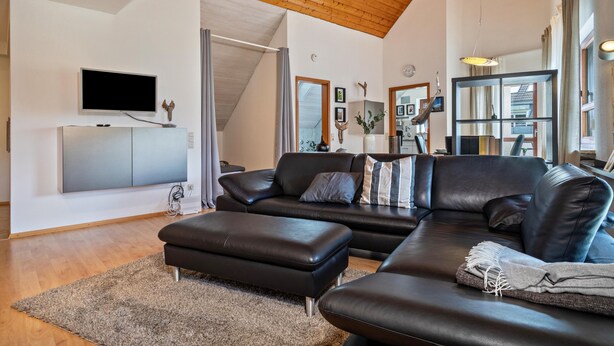 A modern living room with a large black leather sofa, a flat-screen TV mounted on the wall, a wooden ceiling, and a large window. - Retreat Apartment with Mountain View, Balcony, and Wi-Fi (Langenargen)
