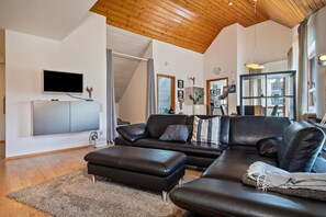 A modern living room with a large black leather sofa, a flat-screen TV mounted on the wall, a wooden ceiling, and a large window.