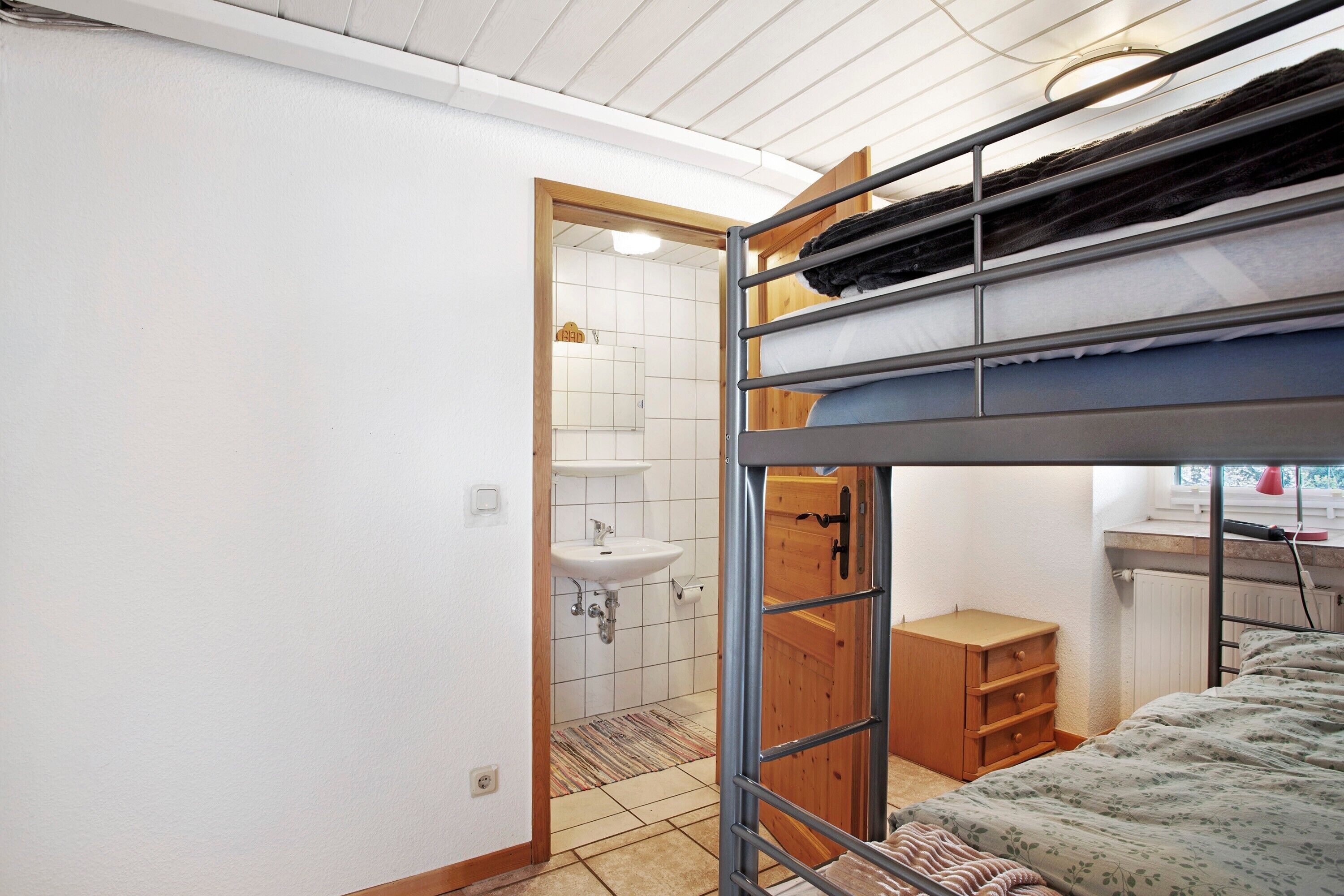 Habitación con literas, una puerta de madera, un pequeño aparador de madera y un lavamanos blanco en el baño contiguo.