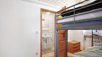 Habitación con literas, una puerta de madera, un pequeño aparador de madera y un lavamanos blanco en el baño contiguo.