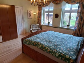 A bedroom with a wooden bed, a chair, a window with curtains, a radiator, and a door.