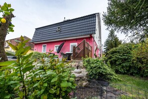 Une maison rose avec un toit noir, un escalier en bois et un jardin avec des plantes et une clôture.