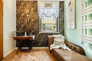Une pièce décorée dans un style rétro, avec une machine à coudre, une horloge ancienne, une fenêtre agrémentée d’un rideau blanc, un canapé marron et un tableau accroché au mur.