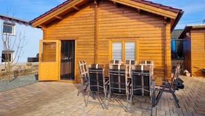 Una cabaña de madera con techo rojo, un patio con sillas de metal y una puerta que da al interior.