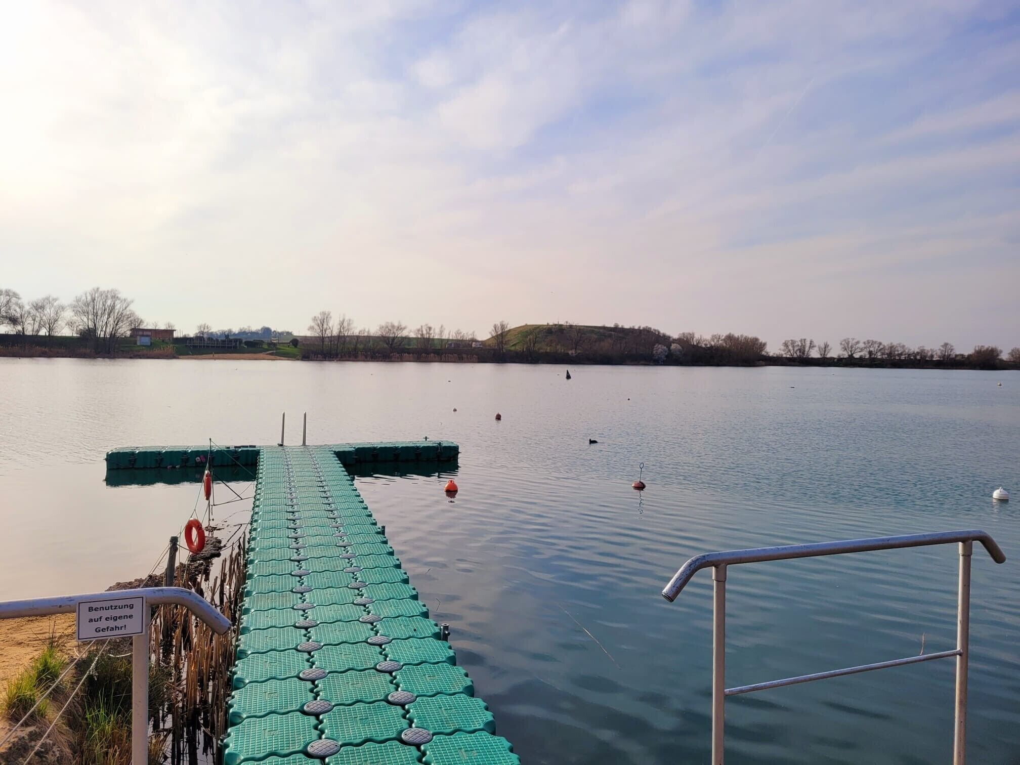A calm lake with a green floating dock extending into the water, surrounded by trees and a distant hill.