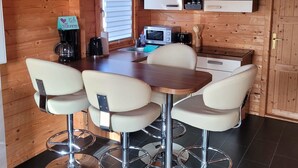 Un interior de madera con comedor amoblado con cuatro sillas y una mesa, una kitchenette con gabinetes y electrodomésticos, y un ventanal con persianas.