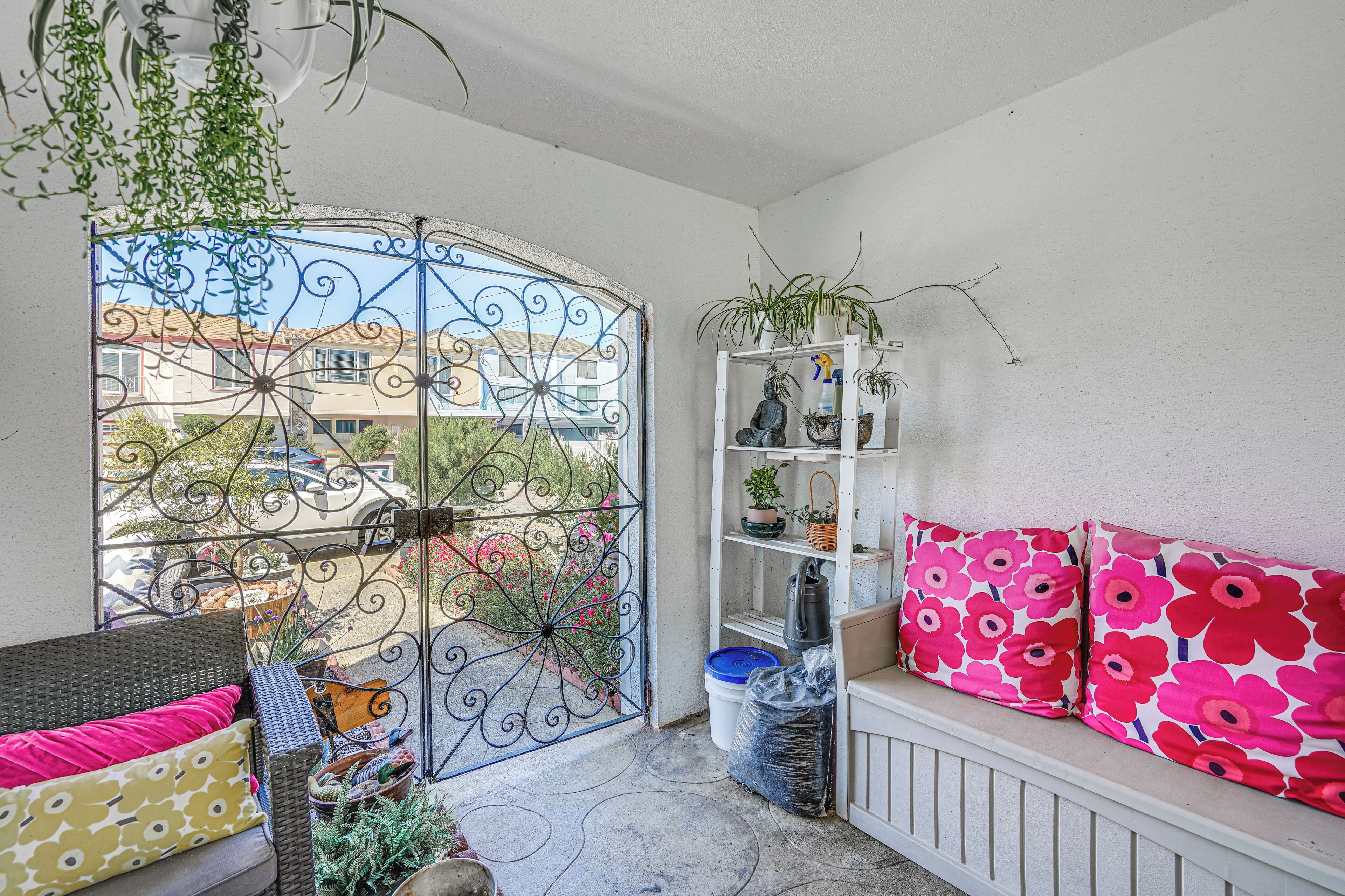 A balcony with a wrought iron gate, a white bench with pink floral cushions, a white shelving unit with plants, and a wicker sofa with patterned cushions.