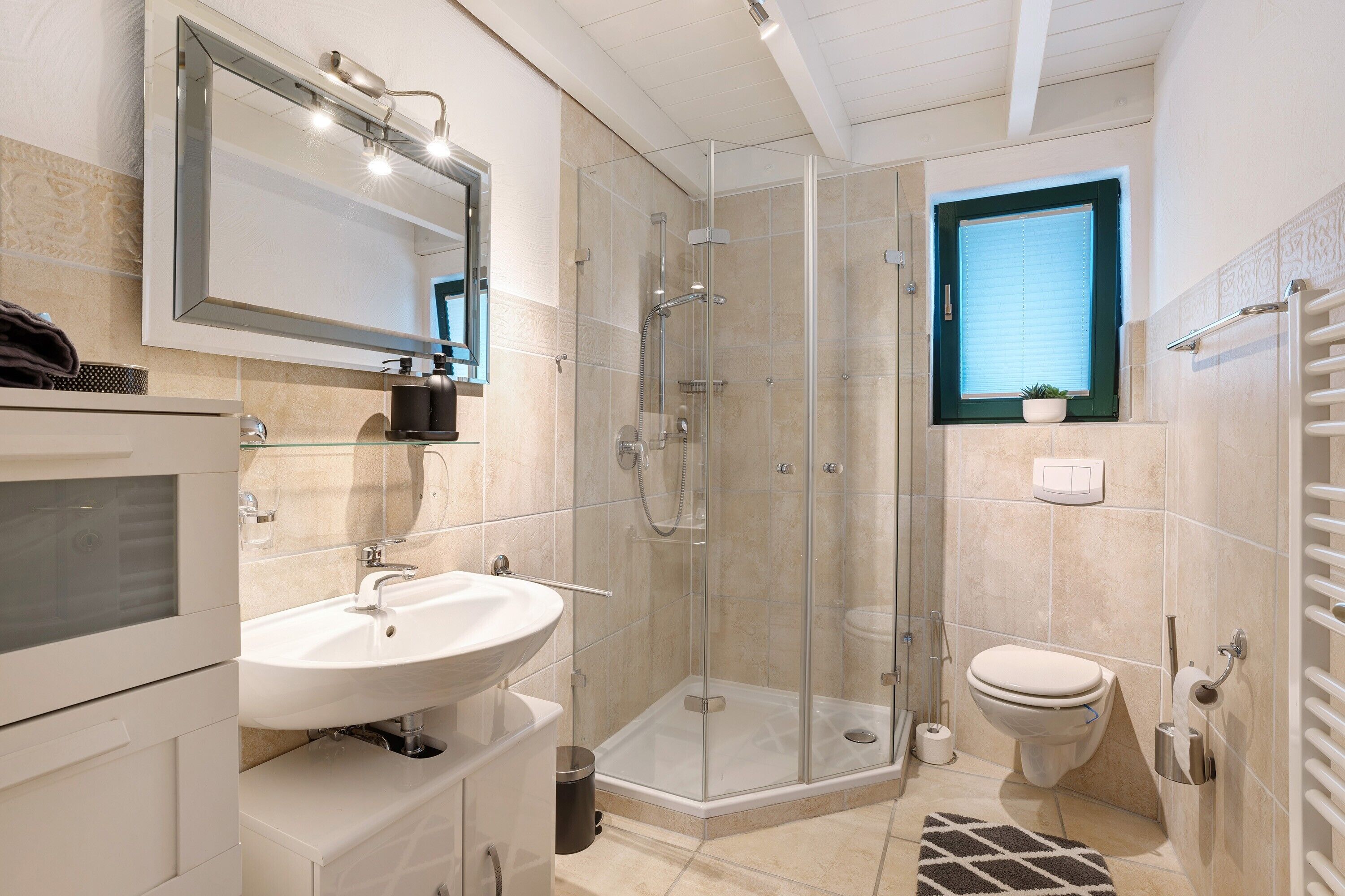 A modern bathroom with a glass shower enclosure, a white sink, a toilet, and a window with blinds.