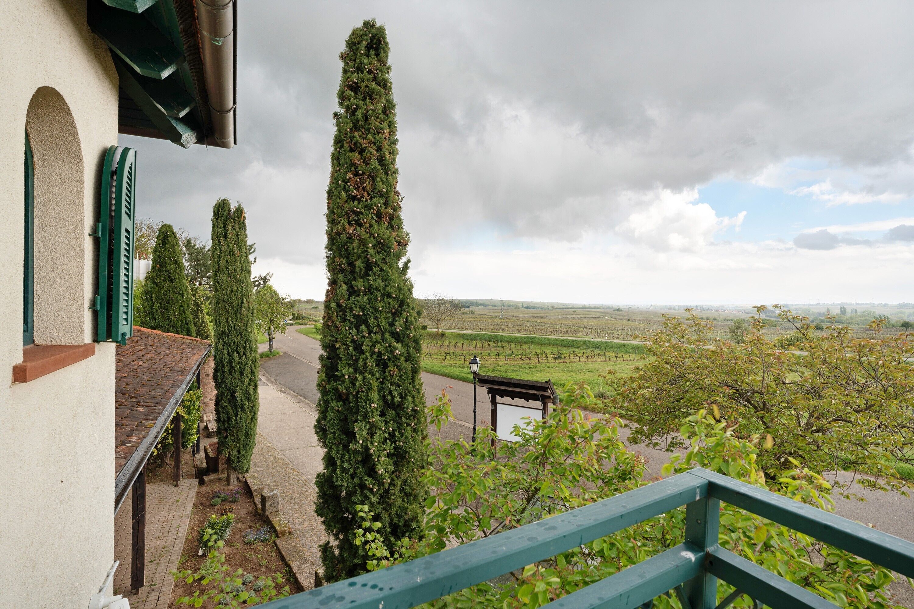 A balcony with a view of a vineyard, a tall tree, and a building with a tiled roof.