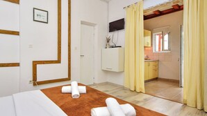 A hotel room with a bed, a brown towel, and a white towel on a brown rug. There is a TV mounted on the wall, a door leading to a kitchen, and a window with curtains. - Nice apartment for 3 people with WIFI, A/C, TV and terrace (Zadar)