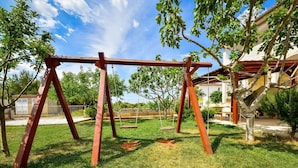 Une aire de jeux avec des balançoires et un espace délimité par une clôture, entouré d’herbe et d’arbres.
