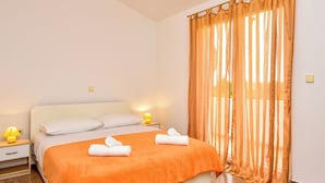 Un lit bien fait, recouvert d’une couvre-lit orange, avec deux oreillers et deux serviettes enroulées. On y trouve une table de chevet avec une lampe, une fenêtre ornée de rideaux orange et un plancher en bois.