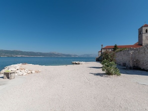A coastal building with a stone wall and a red-tiled roof, adjacent to a pebble beach and clear blue sea. - Nice apartment for 5 guests with WIFI, A/C, TV and terrace (Solin)
