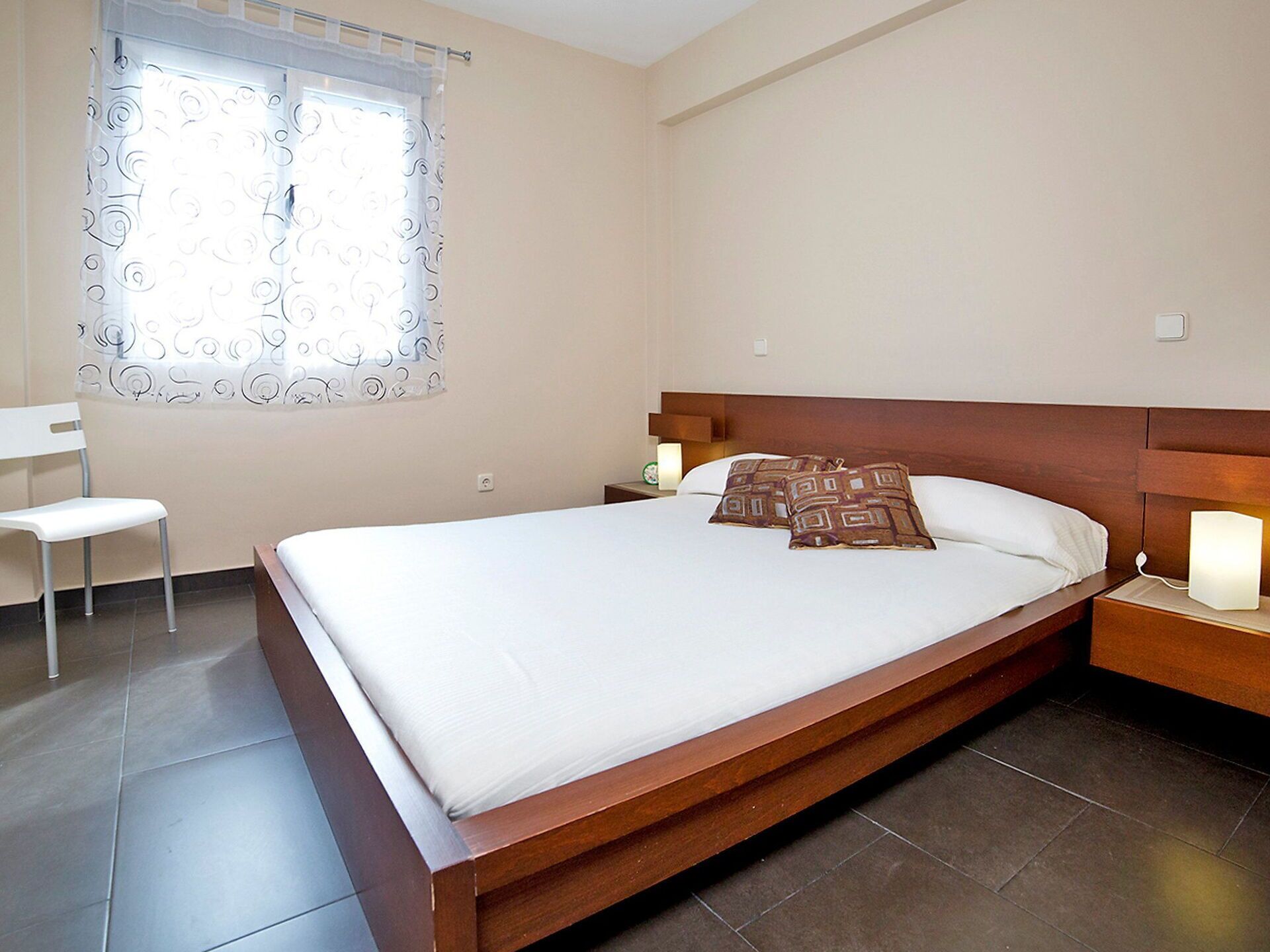 A neatly made bed with a wooden headboard, a bedside table with a lamp, a chair, and a window with patterned curtains.