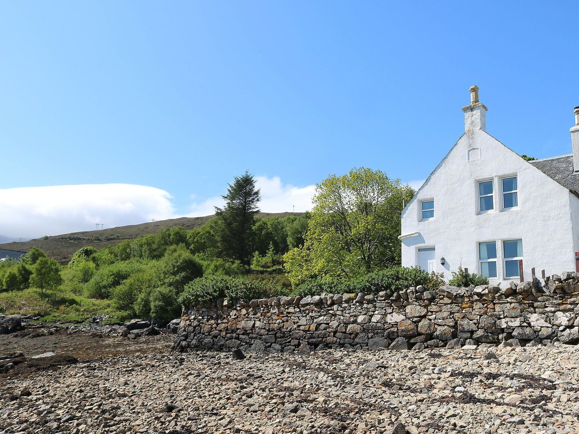 Une maison blanche avec un mur de pierre à l’avant, entourée de verdure et sous un ciel dégagé.