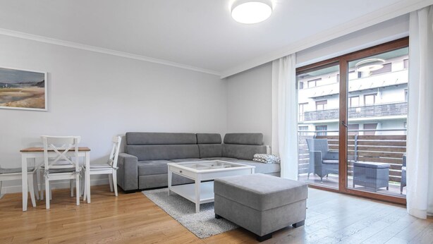 Ein modernes Wohnzimmer mit grauem Sofa, weißem Couchtisch und passendem Hocker. Ein Essbereich mit weißen Stühlen und einem kleinen Tisch ist ebenfalls vorhanden. Der Raum verfügt über Holzdielen und eine große Schiebeglasdoor, die auf einen Außenbalkon mit Korbmöbeln führt.