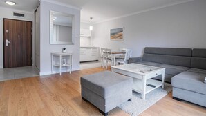 Ein modernes Wohnzimmer mit grauem Sofa, grauem Hocker und weißem Couchtisch. Ein Essbereich mit weißen Stühlen und einem Tisch, sowie eine Küche mit weißen Schränken ist ebenfalls vorhanden. Ein großer Spiegel ist an der Wand angebracht und ein Gemälde ist sichtbar.