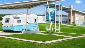 A modern RV parked in a well-maintained grassy area with a covered outdoor seating area and a contemporary building in the background.
