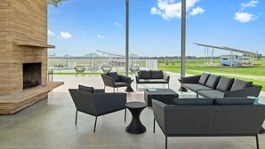 A covered outdoor seating area with modern furniture, a brick fireplace, and a view of a golf course and distant structures.
