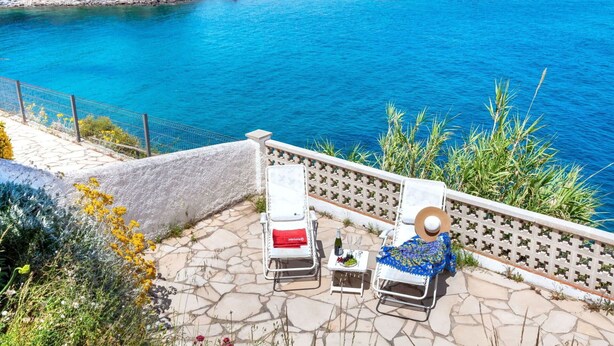 Une terrasse avec deux chaises longues, une table avec une bouteille et des verres, et une vue sur la mer et les montagnes.