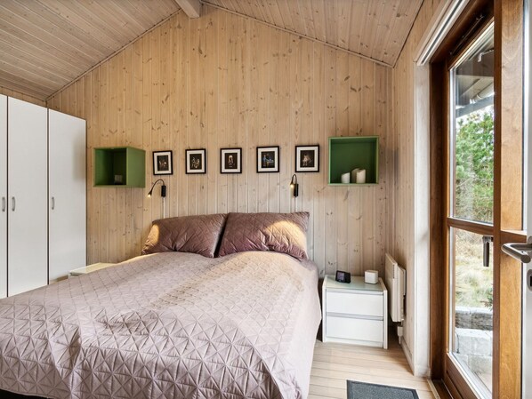 Un dormitorio con techo de madera, cama con cubrecama acolchado, mesita de noche con lámpara y cuadros enmarcados en la pared.