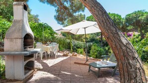 Eine Terrasse im Freien mit einem Steinofen, weißen Stühlen und einem Tisch unter einem großen Sonnenschirm.