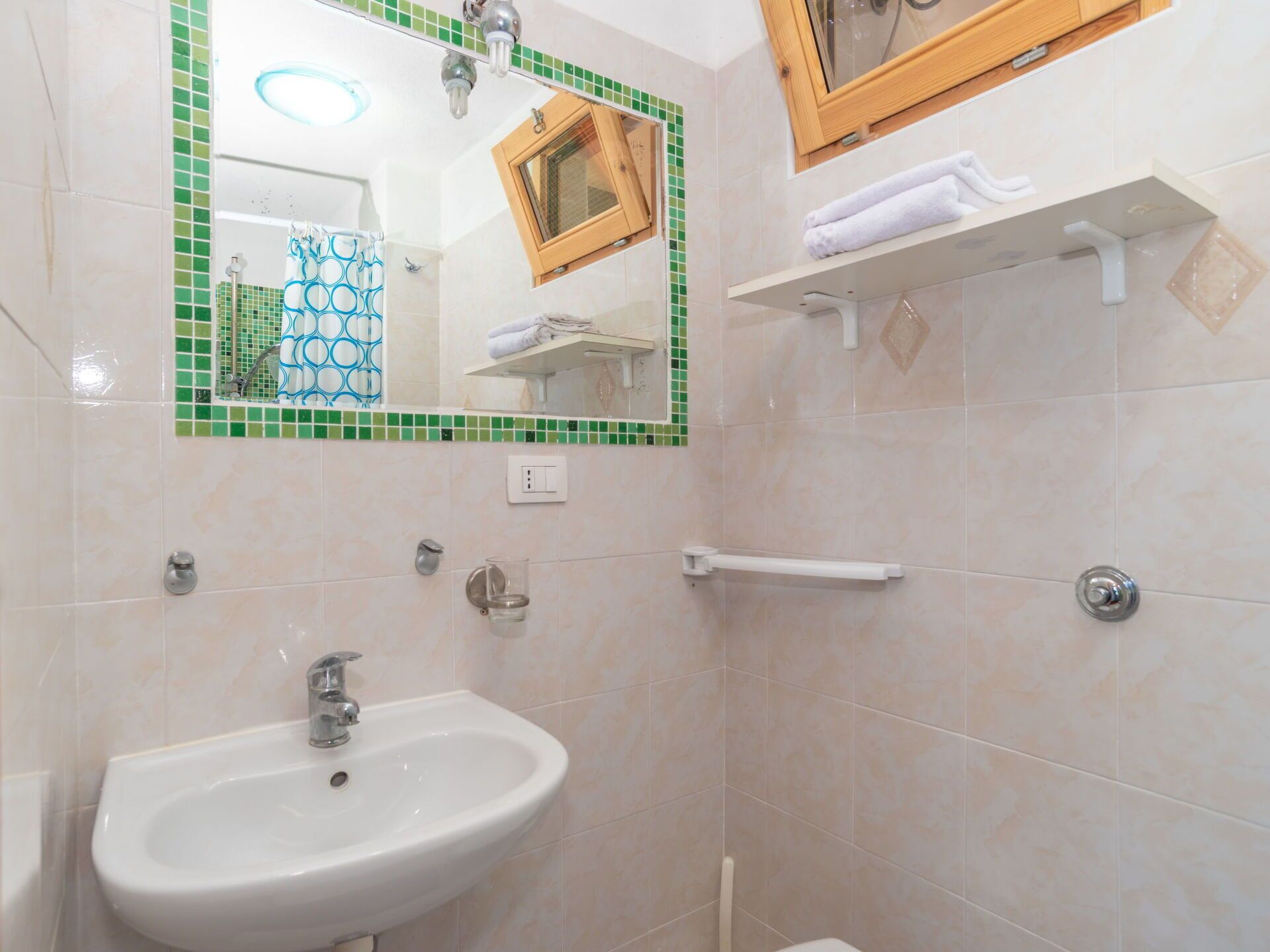 Une salle de bain avec un lavabo blanc, un miroir bordé de carreaux verts, un porte-serviettes et un espace douche.