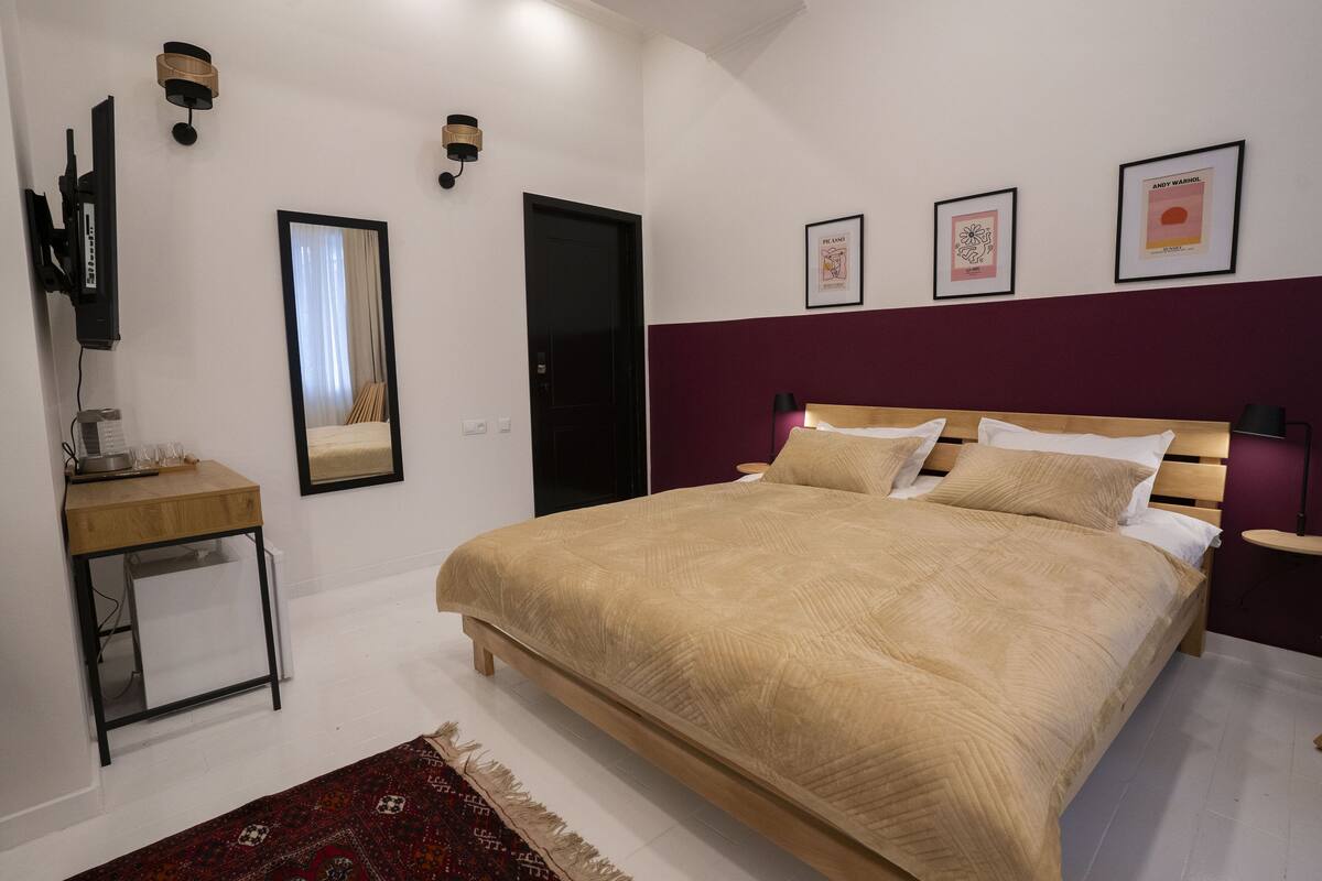A bedroom with a large bed, a bedside table, a mirror, and a wall-mounted TV.