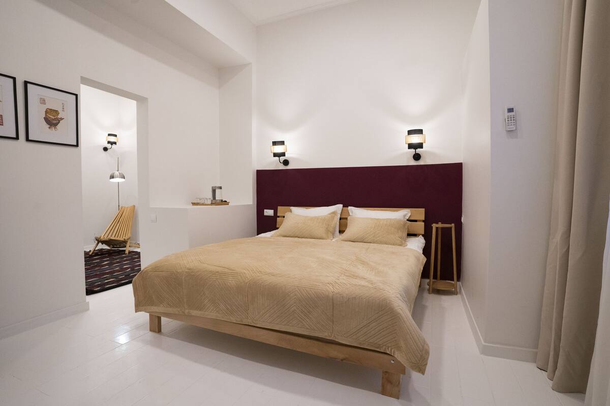 A modern bedroom with a large bed, two bedside tables, wall-mounted lights, a wooden headboard, a chair, and a small table.