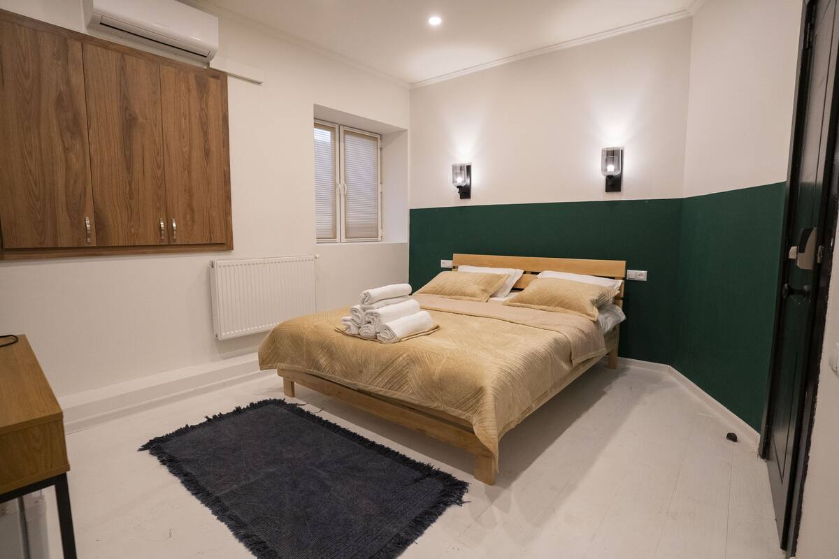 A bedroom with a bed, a wooden wardrobe, a window with blinds, a radiator, and a wall-mounted light fixture.