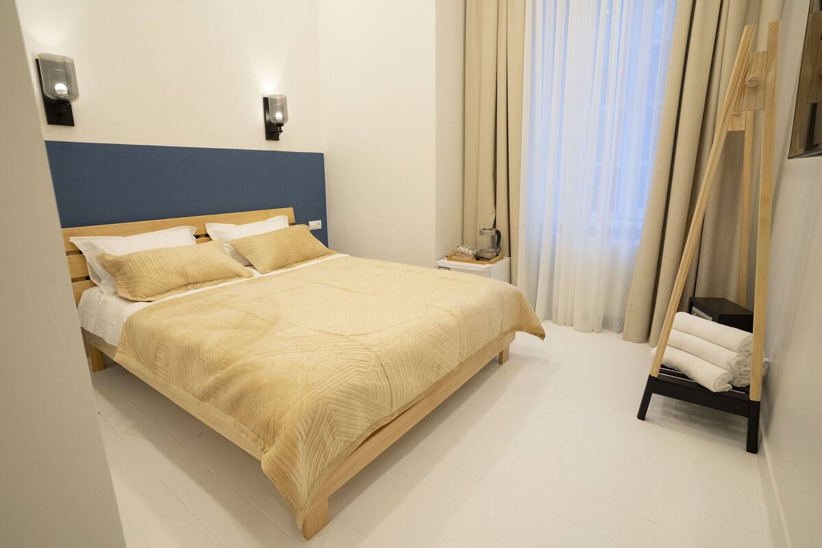 A neatly made bed with a blue headboard, a bedside table with a lamp, and a wooden shelf with towels.