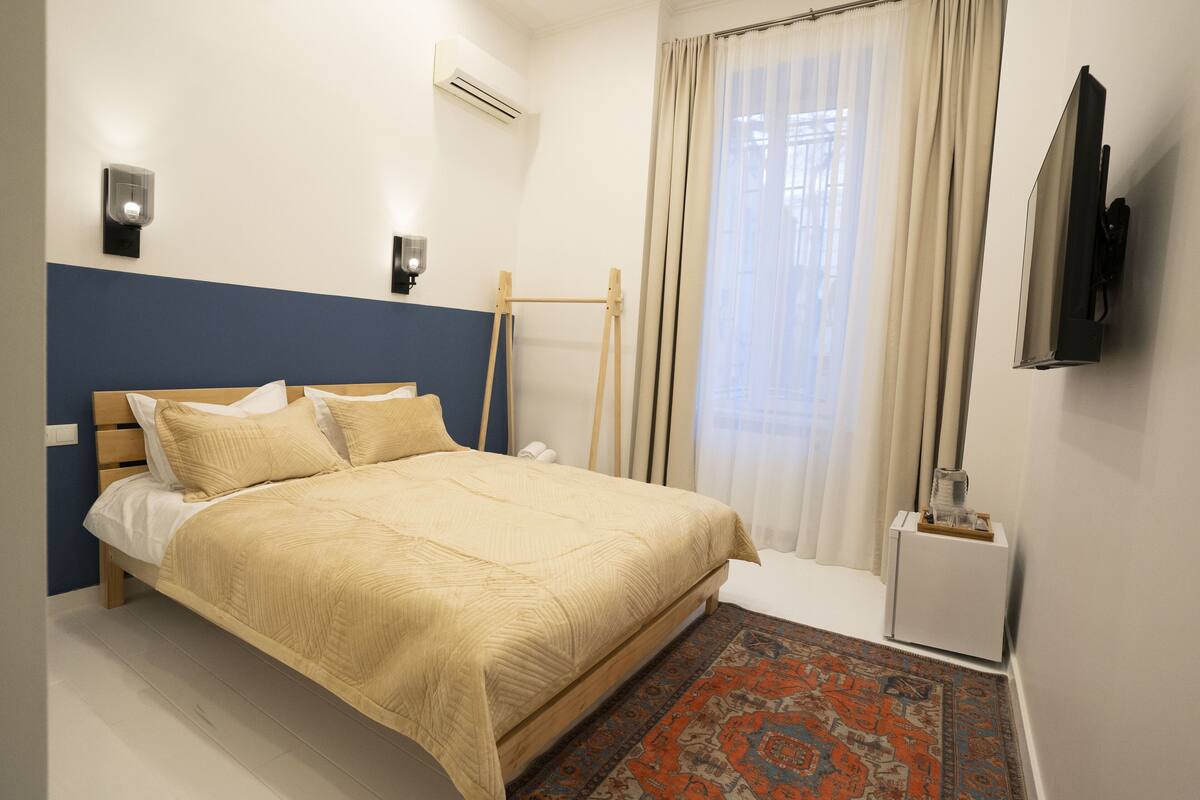 A neatly made bed with a beige comforter and pillows, a wooden headboard, a small bedside table with a lamp, and a wall-mounted TV.
