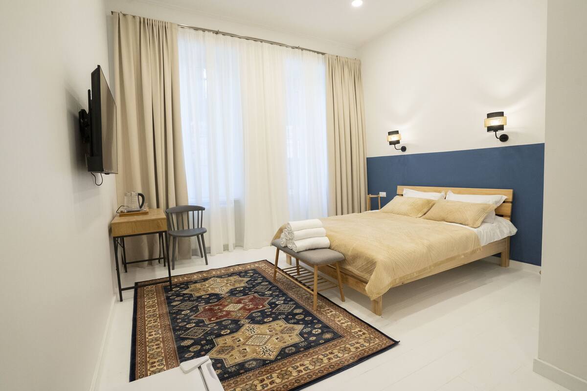 A bedroom with a bed, a desk, a chair, a TV, and a window with curtains.