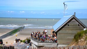 Ein Restaurant direkt am Strand mit Außenbestuhlung mit Blick auf das Meer, ein Holzhaus mit geneigtem Dach und ein Sandstrand im Hintergrund.