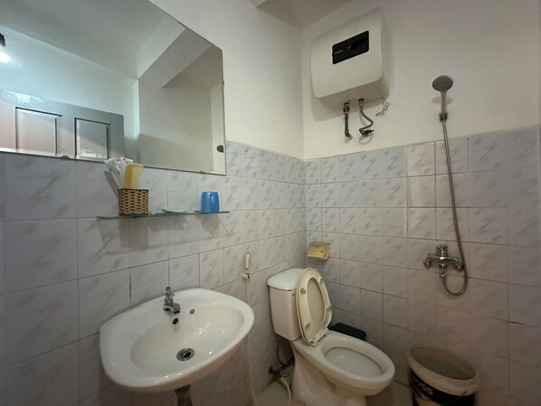 Une salle de bain équipée d’un lavabo, de toilettes et d’une douche. Un miroir se trouve au-dessus du lavabo, et une étagère contient des articles de toilette.
