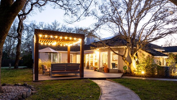 Ein Außenbereich mit Terrasse, einer Pergola, Sitzgelegenheiten und einem Weg zu einem Haus.