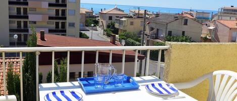 A balcony with a table set for two, overlooking a coastal town with terracotta-roofed buildings.
