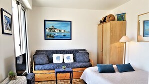 A cozy room with a sofa, a bed, a wooden wardrobe, and a painting of a coastal scene. - Nice apartment for 2 guests with WIFI, TV, balcony and pets allowed (Narbonne)