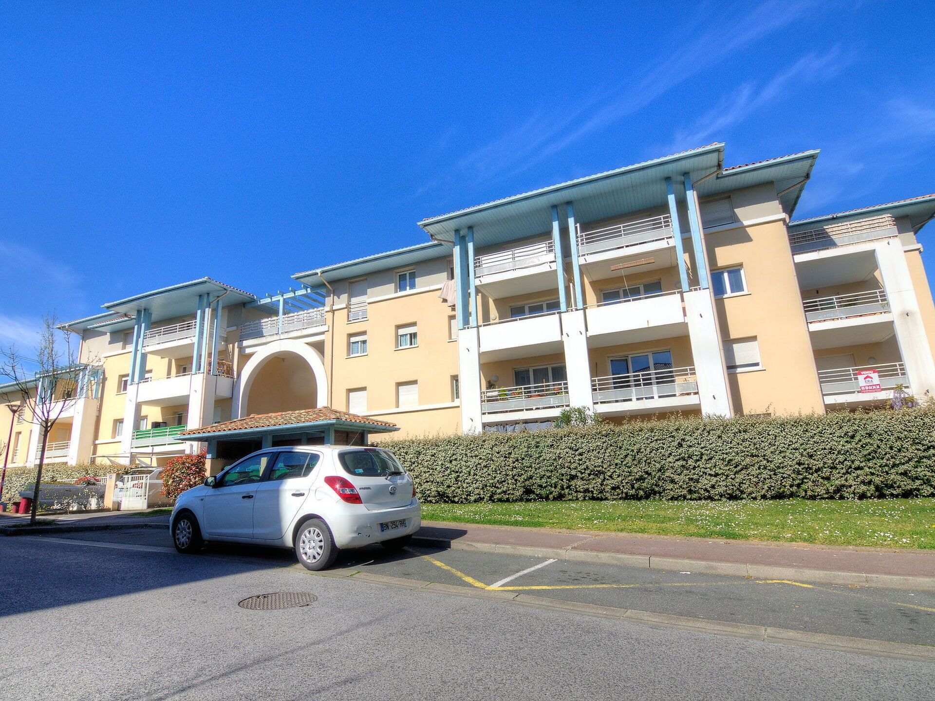A multi-story residential building with balconies, a parking area, and a hedge in front.