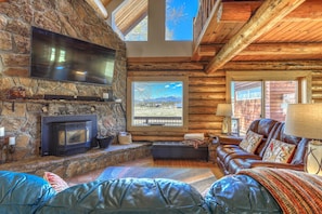 L’intérieur chaleureux d’un chalet en bois, avec une cheminée en pierre, un canapé en cuir et un grand téléviseur fixé au mur.