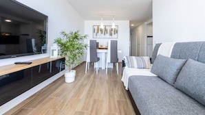 A modern living room with a grey sofa, a dining area with a white table and chairs, and a large flat-screen TV on the left. - Cosy apartment for 5 guests with A/C, WIFI, TV and balcony (Salou)
