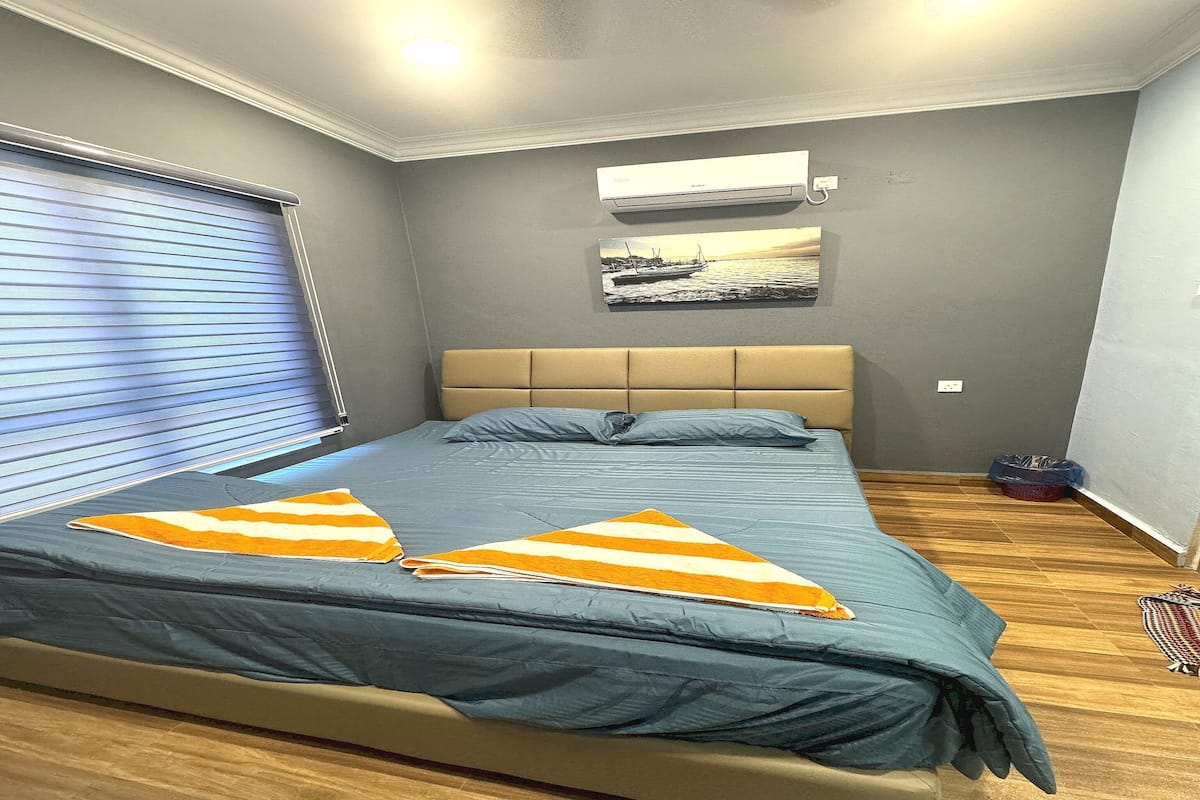 A bedroom with a bed, an air conditioner, a window with blinds, and a picture on the wall.