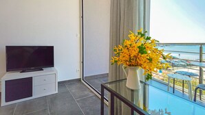 Ein modernes Zimmer mit einem Flachbildfernseher auf einer weißen Kommode, einem Glastisch mit einer Vase gelber Blumen und einem Balkon mit Blick auf das Meer und parkende Autos.