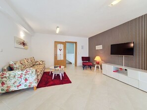 Una sala de estar con un sofá, una butaca roja, una mesita, un televisor y un televisor de pared.