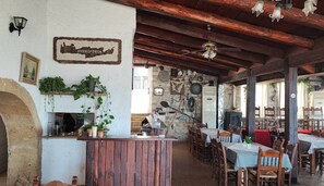 A rustic interior with a stone wall, wooden ceiling beams, a bar area, and tables set for dining. - Glikanisos House Nipos - Ideal for Hikers and Families 2bd, 6pl, 10min to Beach (Apokoronas)