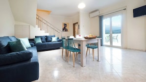 A modern living room with a sofa set, dining table, chairs, and a staircase leading to an upper floor. - Glikanisos House Nipos - Ideal for Hikers and Families 2bd, 6pl, 10min to Beach (Apokoronas)