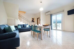A modern living room with a sofa set, dining table, chairs, and a staircase leading to an upper floor. - Glikanisos House Nipos - Ideal for Hikers and Families 2bd, 6pl, 10min to Beach (Apokoronas)
