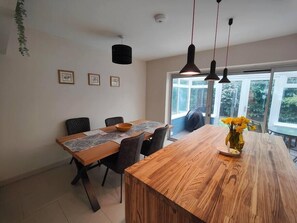 Un coin repas avec une table en bois, des chaises et des lampes suspendues. Les murs sont ornés de tableaux encadrés et une fenêtre offre une vue sur la verdure.