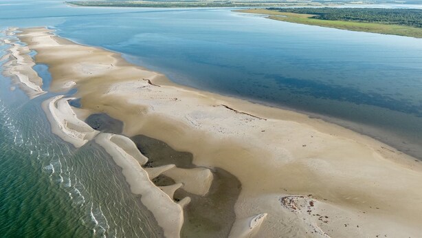Ein Luftbild einer Küstenregion mit Sandstrand, ruhigem Wasser und im Hintergrund Windkraftanlagen.