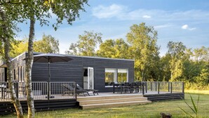 Eine moderne, dunkel gefärbte Holzhütte mit einer Terrasse und Sitzgelegenheiten im Freien, umgeben von Bäumen und Gras.